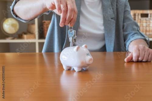 Person's hand placing house keys into a pink piggy bank on a wooden table, symbolizing saving for mortgage, homeownership, real estate investment and future housing expenses