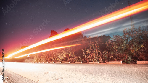 Slika na platnu A starry night sky above a forest road, with vivid red and orange light trails streaking across, capturing motion and speed against the stillness of trees and stars