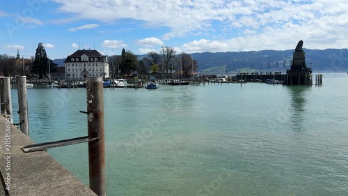 Scenic view of Lindau Harbor on Lake Constance (Bodensee), featuring the historic lighthouse, Bavarian Lion statue, calm waters, and alpine backdrop under a beautiful cloudy sky in southern Germany.