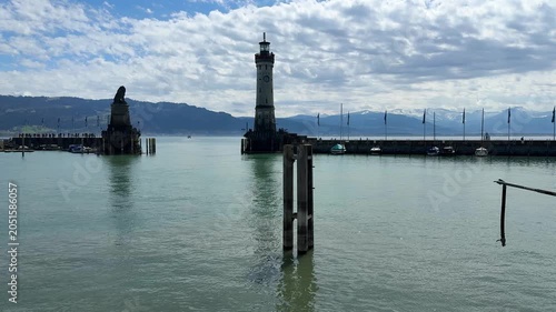 Scenic view of Lindau Harbor on Lake Constance (Bodensee), featuring the historic lighthouse, Bavarian Lion statue, calm waters, and alpine backdrop under a beautiful cloudy sky in southern Germany.