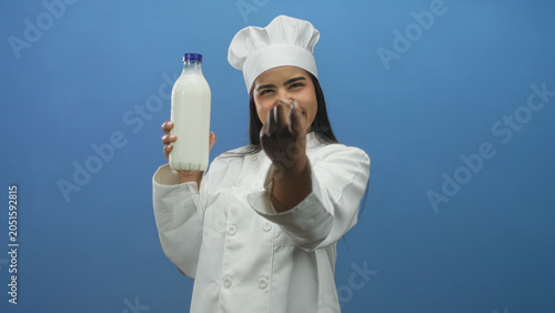 Φωτογραφία Young woman chef smiling holds milk bottle and beckons with fingers in studio; fresh dairy inviting