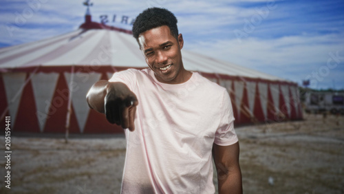Foto Black man pointing finger at camera showing bare hand and smile, circus tent behind on a street fairground under blue sky; confidence invitation