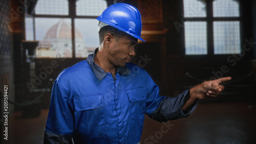 Foto Man in blue hardhat and coveralls points finger toward exhibit inside a museum building with tall arched windows and rope stanchions visible; duty