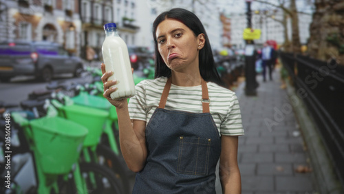 Photography Woman young in apron holds milk bottle in raised hand, pouting lower lip while looking skeptical on a busy city street with parked rental bikes; disappointment