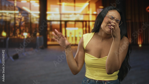 Foto Woman pinches nose outside building entrance wearing yellow top and glasses; odor discomfort disgust