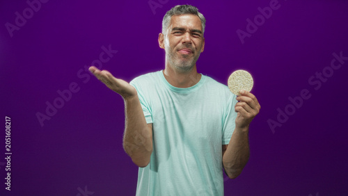 фотографія Man holding a round corn pastry in one hand and pinching his nose with the other in a purple studio set, face cringing at the smell; disgust