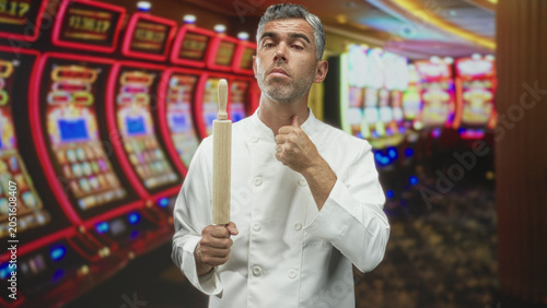 Bild auf Leinwand Chef man holds rollingpin, gives thumbsup and grips utensil amid slot machines in a building casino setting with neon lights and carpeted floor; confidence culinary gamble