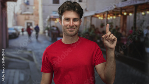 Quadro su tela Man points finger upward on a street terrace outside a restaurant wearing a red t shirt; confidence casual charm