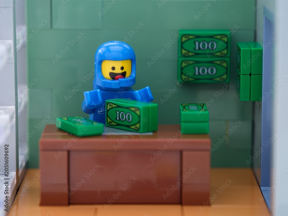 Obraz premium Tambov, Russian Federation - May 26, 2025 A blue Lego astronaut minifigure counts green toy cash bundles at a brown wooden desk.