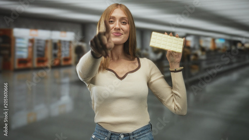 Photos Young redhead woman holds white chocolate bar, smiles and points finger to camera in airport terminal concourse with vending machines; playful invitation