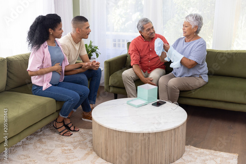 Diverse family with senior couple showing blue onesie at home, pregnant partner in maternity top