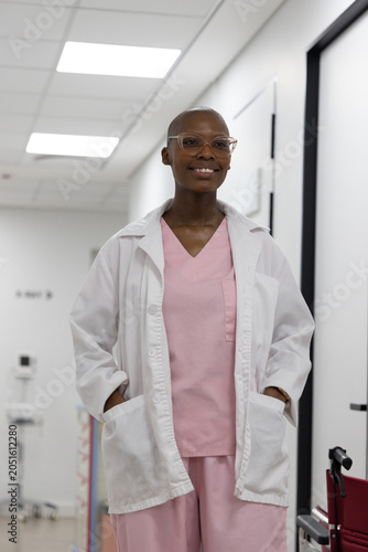 African woman standing in clinic hall wearing pink scrubs lab coat hands in pockets red wheelchair