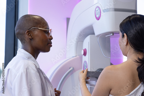 Diverse female tech assisting female patient in back-open gown placing breast on paddle in clinic