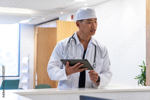 Asian male physician wearing lab coat and stethoscope scrub cap holding tablet at clinic reception