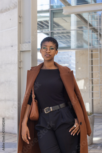 African woman standing at urban plaza wearing brown coat, leather shoulder bag, round eyeglasses