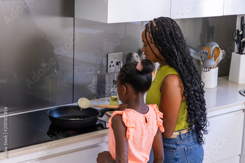 African American mother and teenage daughter cooking at stovetop at home with frying pan, ladle
