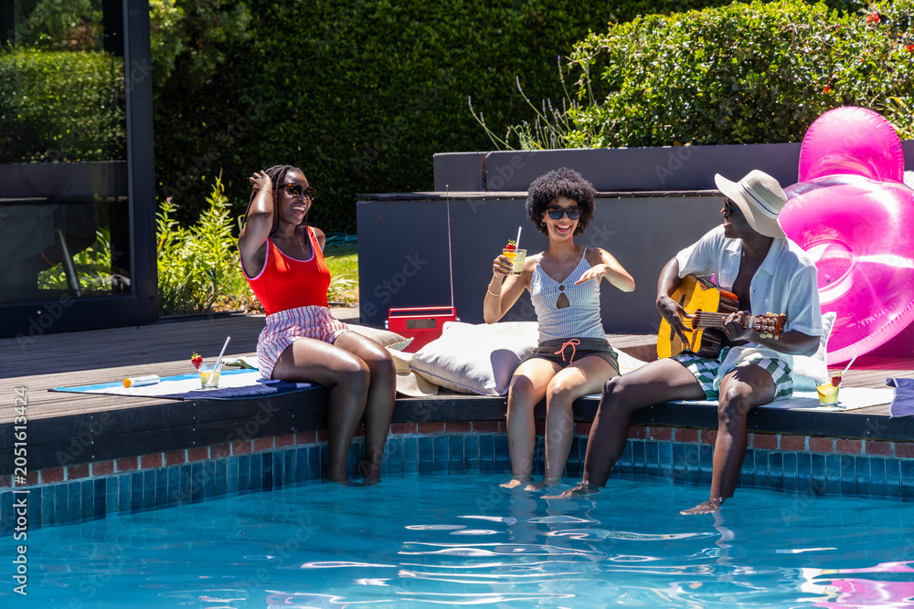 Naklejka premium African American friends sitting on wooden deck at pool, in swimsuits playing guitar, holding drink