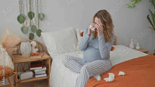Schilderij op canvas Pregnant woman crying and wiping tears with a tissue, hand on belly on a bed in a bedroom with tissues scattered on blanket; sadness