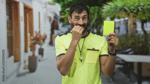 Fotografia Man referee holding yellow card, finger to mouth gesture on street lined with white buildings; authority caution