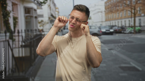 Foto Man posing with fists near his face and smiling on a street in front of parked vehicles and townhouses, wearing glasses and a beige t shirt; playful confidence