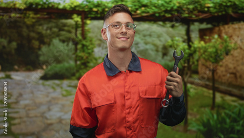Fotografia Man mechanic in orange coverall holding wrench in right hand, smiling and looking up beside stone path under pergola in a building courtyard; confidence craft