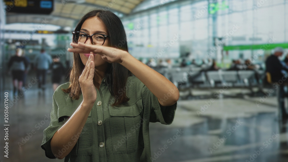 Fototapeta premium Woman with hands in timeout gesture at airport terminal wearing glasses and olive green shirt, facing camera; delay assertion.