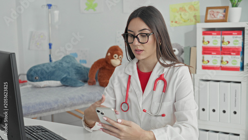 Valokuva Woman doctor in white coat with red stethoscope holding smartphone, right hand touching screen, seated at computer desk with plush toys on exam bed in building; focused care