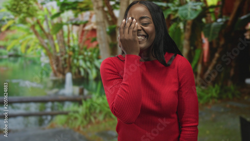 Photos Woman in red sweater covers face with hand by a garden pond fountain, smiling and glancing upward; shyness joy