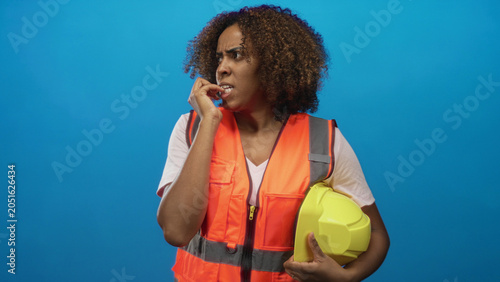 Εκτύπωση καμβά Woman engineer african american wearing orange safety vest holding yellow hard hat and biting nails in blue studio; nervous concern