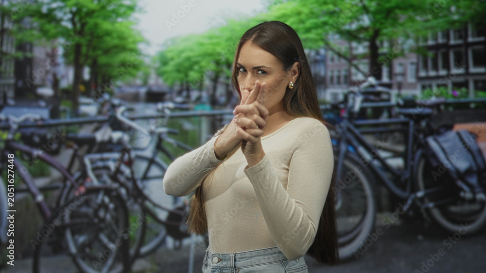 Fototapeta premium Woman forming finger gun with hands and pursed lips, visible fingers aimed forward on a street by bicycles and canal; playful confidence.