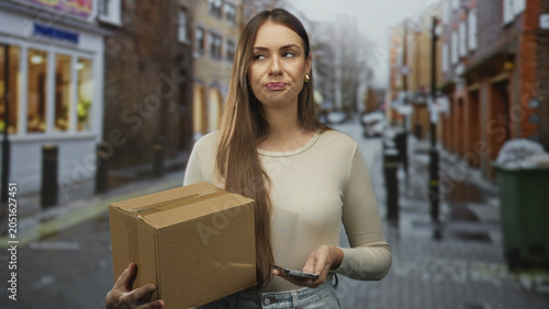 Φωτογραφία Woman holds cardboard box and taps phone while standing on street, expression flat and unimpressed; annoyance waiting