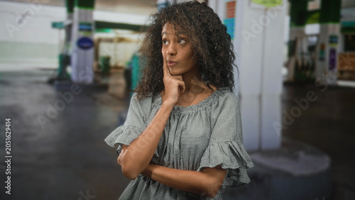 Fotografija Teen woman with curly hair touching her cheek with index finger, arms crossed while standing by a street gas pump; thoughtful doubt decision