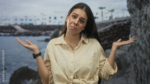 Foto Woman shrugging with palms up and tilted head on a street by the seaside, light shirt knotted at waist and smartwatch visible; uncertainty