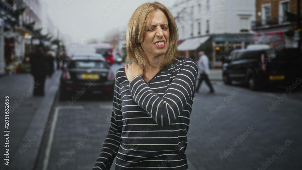 Fototapeta premium Woman clutching shoulder with hand, wincing in pain on a street lined with parked cars and shops; discomfort strain.