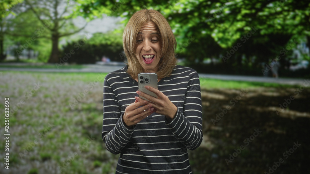 Fototapeta premium Woman holding smartphone, tapping screen with hands and smiling while reading messages in forest sunlight; joy connection.