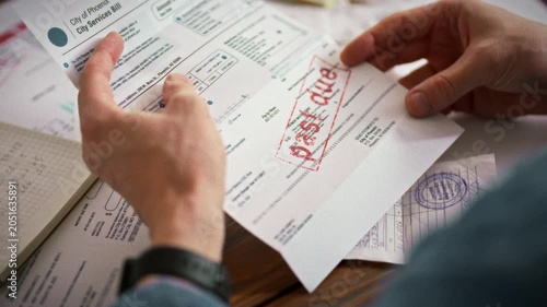A man scrutinizes a receipt, pulled from an envelope marked overdue, against a backdrop of unpaid bills, highlighting the stress of managing late utility payments and outstanding debts