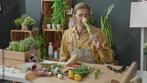 Foto Man florist smelling a yellow tulip at studio table with apron, pruners and vases and plants; calm creativity