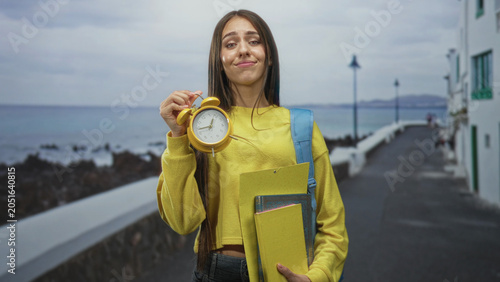 Fotografie Teenage student holds a yellow alarm clock and school folders on a street by the sea, lifting the clock with a smirk and raised wrist gesture; time pressure playful