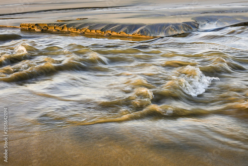 Stream flows across sandy beach cutting path with waves of erosion