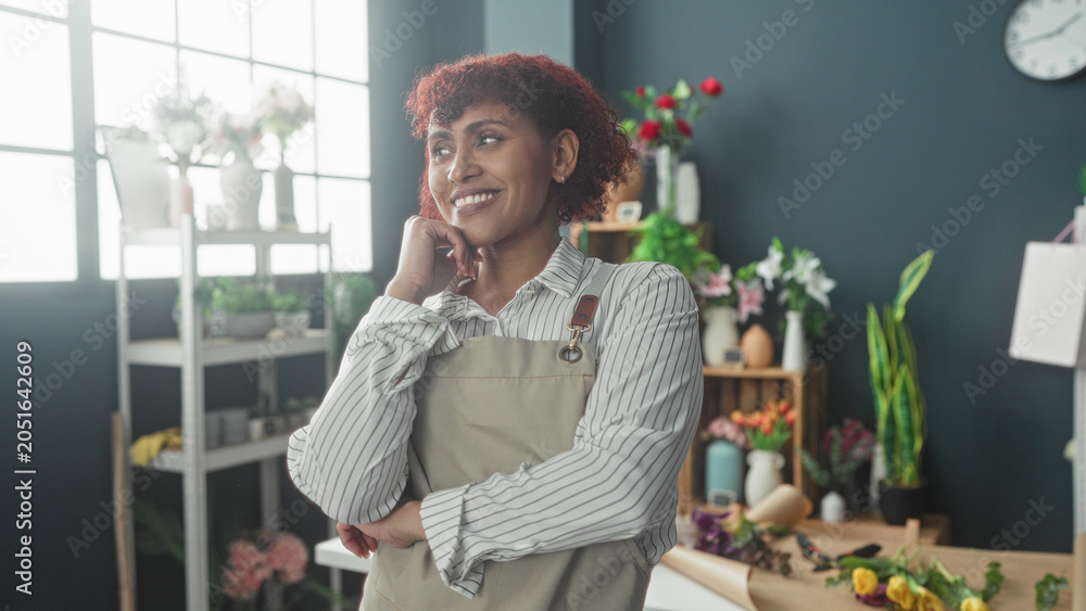 Fototapeta premium Woman florist in apron hands on hips beside bouquets and seedlings on a worktable in studio; quiet contentment.