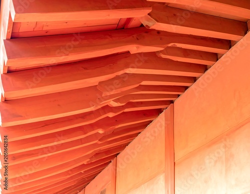 Close-up of a wooden structure's roof. Horizontal beams are visible, with intricate carved details and rich, warm colors. The angled ceiling suggests an overhang