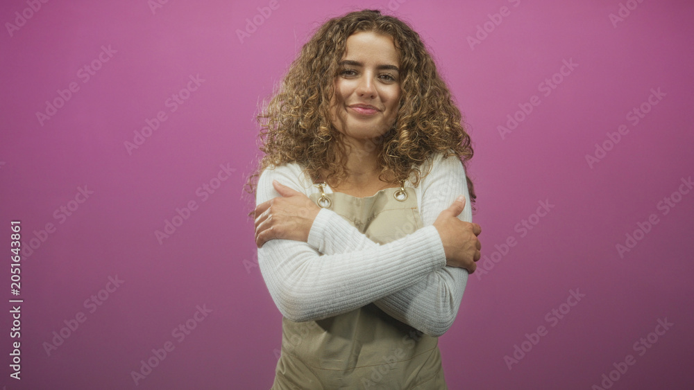 Fototapeta premium Young woman wearing apron hugs herself with crossed arms and visible hands in a studio against a pink backdrop; comfort serenity.