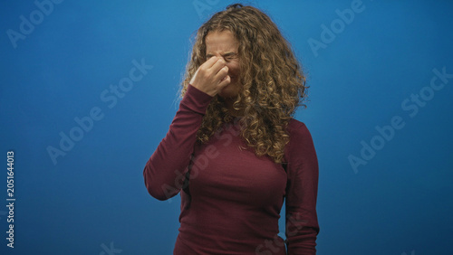 Obraz na plátně Woman with curly hair wearing a maroon sweater pinches nose with fingers and closes eyes in studio blue backdrop; disgust