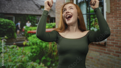 Schilderij op canvas Redhead woman pointing both index fingers upward with bare shoulders visible, smiling in front of a brick building and lush garden; joy celebration