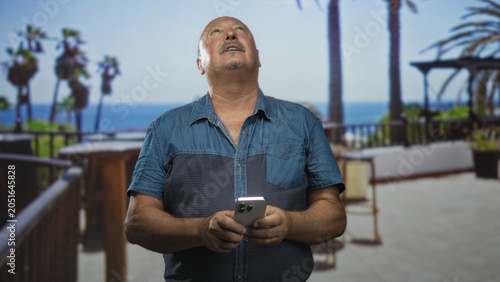 Canvas-taulu Man texting on smartphone on oceanfront terrace holding phone with both hands and glancing down at screen; calm reflection