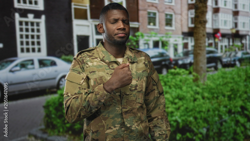 Man in camouflage soldier uniform points to chest with hand on street in front of building and parked cars; pride duty.