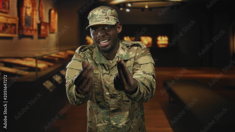 Fototapeta premium Soldier man in camouflage uniform extends palms forward in museum building with display cases and framed art; pride duty.