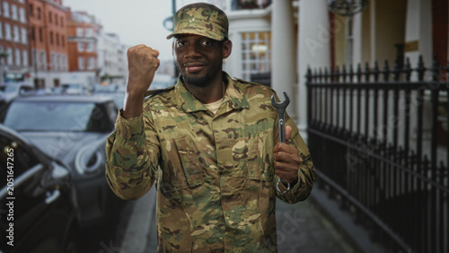 Fotografía Man in camouflage uniform holding wrench and raising fist on street beside cars and iron railing; pride duty resilience