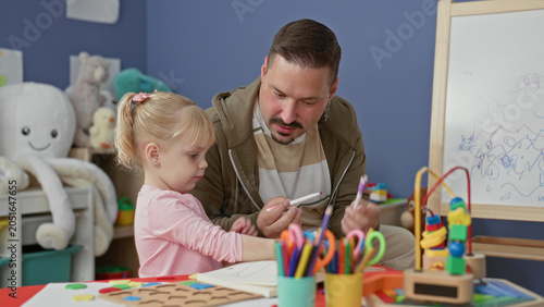 Obraz na plátně Man guiding young girl to hold markers and color on paper at a preschool building while seated at a craft table strewn with toys and pencils; learning patience