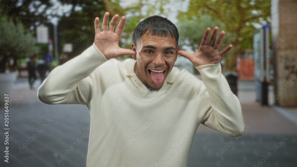 Naklejka premium Young hispanic man sticks out tongue and raises hands to temples in a goofy facial gesture on street, smiling at camera in a cream sweater and trimmed goatee; playful joy.